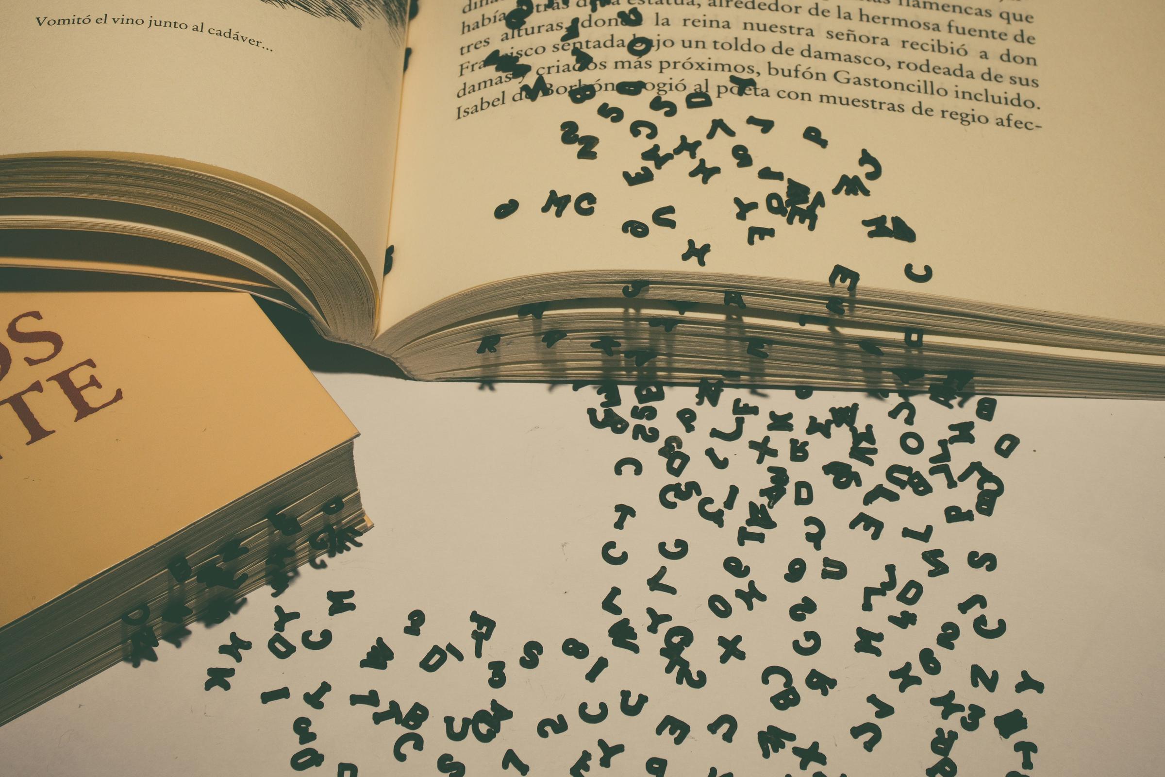 Small cut-out letters spilling from the open pages of a book onto a pale table.